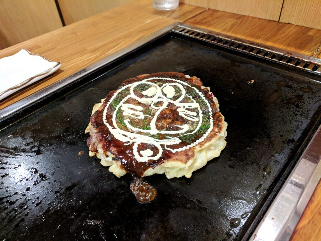 Osaka, Japan Foodie Travelogue - Okaru restaurant serving Okonomiyaki and Yakisoba. Booth seat in the restaurant, with grill at table. Seen here is the okonomiyaki with Doraemon's face drawn with mayonaise.